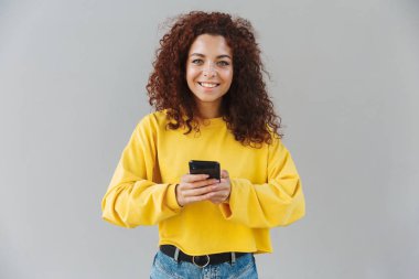 Mutlu neşeli güzel kıvırcık kız cep telefonu kullanarak gri arka plan üzerinde izole.
