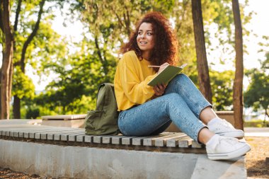 Genç güzel kıvırcık öğrenci kız dış mekanda doğa parkı not notları yazma oturan.