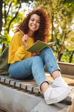 Genç güzel kıvırcık öğrenci kız dış mekanda doğa parkı not notları yazma oturan.