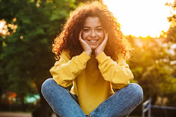 Erfreut glücklich niedliche junge Studentin lockiges Mädchen sitzt auf Bank im Freien im Naturpark mit schönem Sonnenlicht. — Stockfoto