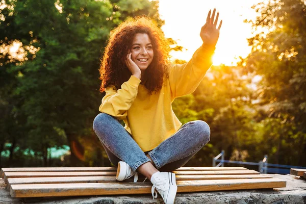 Feliz lindo joven estudiante rizado chica sentada en el banco al aire libre en el parque natural con hermosa luz del sol mirando a un lado saludando a los amigos . —  Fotos de Stock
