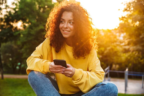 Feliz optimista lindo estudiante rizado chica sentada en el banco al aire libre en el parque natural con hermosa luz del sol utilizando el teléfono móvil . —  Fotos de Stock