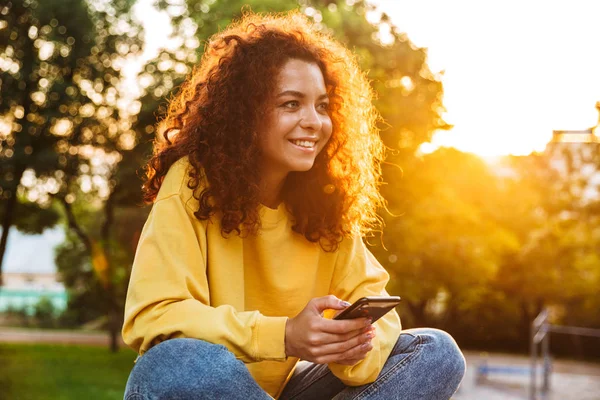 Feliz alegre linda estudiante rizado chica sentada en el banco al aire libre en el parque natural con hermosa luz del sol utilizando el teléfono móvil . —  Fotos de Stock