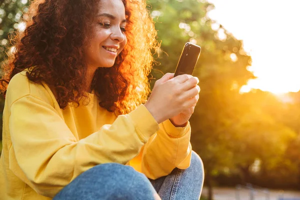 Feliz sorrindo bonito jovem estudante encaracolado menina sentado no banco ao ar livre no parque da natureza com bela luz solar usando telefone móvel . — Fotografia de Stock