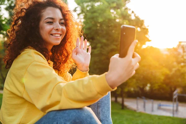 Ευτυχισμένο χαρούμενο χαριτωμένο νεαρό μαθητή σγουρά κορίτσι κάθεται στο πάγκο σε εξωτερικούς χώρους στο φυσικό πάρκο με όμορφο ηλιακό φως χρησιμοποιώντας το κινητό τηλέφωνο. — Φωτογραφία Αρχείου