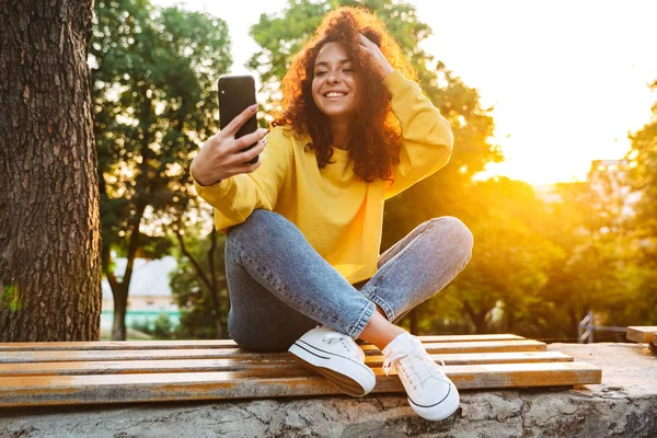 Feliz optimista lindo estudiante rizado chica sentada en el banco al aire libre en el parque natural con hermosa luz del sol utilizando el teléfono móvil . —  Fotos de Stock