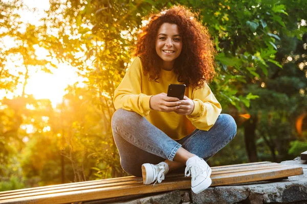Alegre sonriente lindo estudiante rizado chica sentada en el banco al aire libre en el parque natural con hermosa luz del sol utilizando el teléfono móvil . —  Fotos de Stock