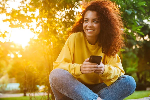 Imagem de uma jovem mulher bonita sorrindo e segurando smartphone enquanto se senta no banco no parque verde — Fotografia de Stock