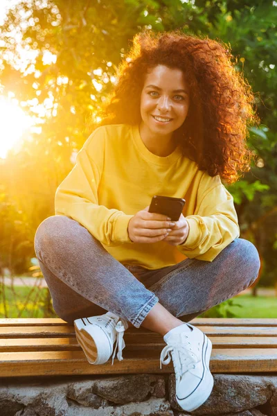 Imagen de la joven caucásica sonriendo y sosteniendo el teléfono inteligente mientras está sentada en el banco en el parque verde —  Fotos de Stock