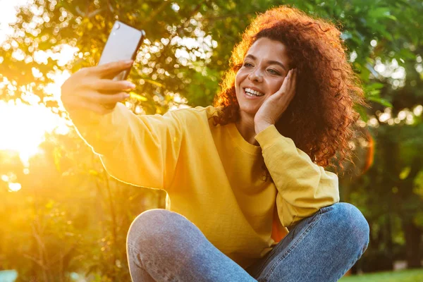 Imagen de una joven encantadora sonriendo y tomando una foto selfie en el teléfono inteligente mientras está sentada en el banco en el parque verde —  Fotos de Stock