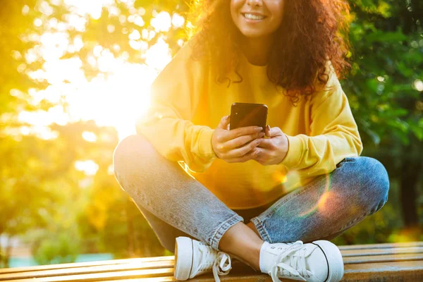 Imagen recortada de la encantadora mujer joven sonriendo y sosteniendo el teléfono inteligente mientras está sentado en el banco en el parque verde —  Fotos de Stock