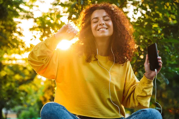 Imagen de una joven feliz escuchando música con smartphone y auriculares mientras está sentada en el banco en el parque verde —  Fotos de Stock