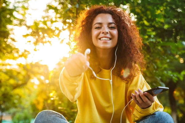 Imagen de una mujer joven y bonita escuchando música con smartphone y auriculares mientras está sentada en el banco en el parque verde —  Fotos de Stock