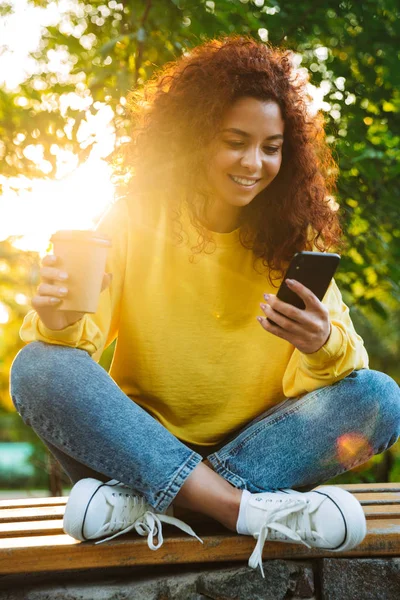 Feliz alegre sonrisa optimista lindo estudiante rizado chica sentada en el banco al aire libre en el parque natural con hermosa luz del sol utilizando el teléfono móvil . —  Fotos de Stock