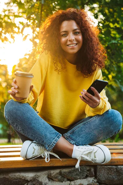 Feliz optimista lindo estudiante rizado chica sentada en el banco al aire libre en el parque natural con hermosa luz del sol utilizando el teléfono móvil . —  Fotos de Stock