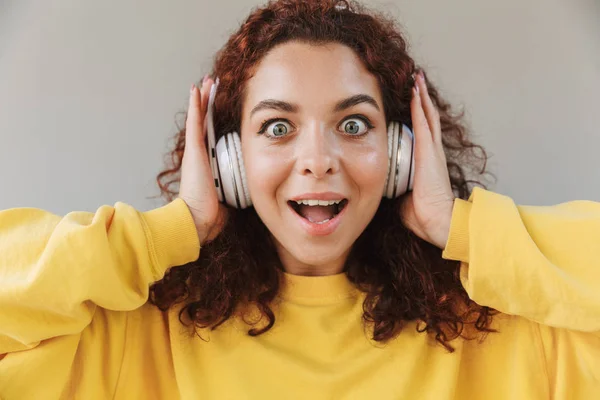 Σοκαρισμένος ενθουσιασμένος όμορφη σγουρά κορίτσι απομονώνεται πάνω από γκρι φόντο ακούγοντας μουσική με ακουστικά. — Φωτογραφία Αρχείου
