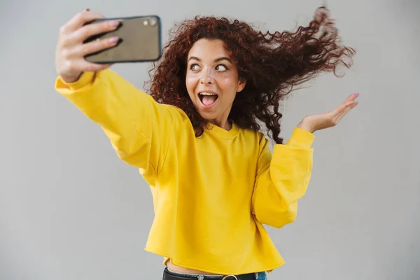 Emocionado chocado linda garota encaracolado isolado sobre fundo cinza usando telefone celular tirar uma selfie . — Fotografia de Stock