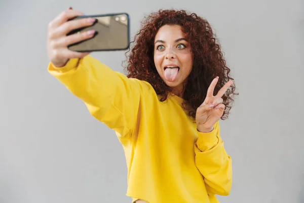 Glücklich fröhlich schöne lockige Mädchen isoliert über grauen Hintergrund mit Handy ein Selfie machen. — Stockfoto
