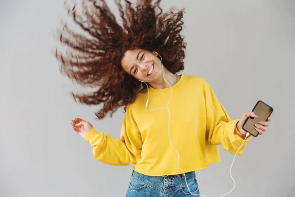 Όμορφη σγουρά κορίτσι απομονώνεται πάνω από το γκρι φόντο χρησιμοποιώντας κινητό τηλέφωνο ακούγοντας μουσική κουνώντας τα μαλλιά — Φωτογραφία Αρχείου