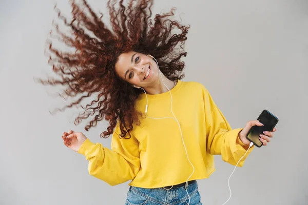 Όμορφη σγουρά κορίτσι απομονώνεται πάνω από το γκρι φόντο χρησιμοποιώντας κινητό τηλέφωνο ακούγοντας μουσική κουνώντας τα μαλλιά — Φωτογραφία Αρχείου