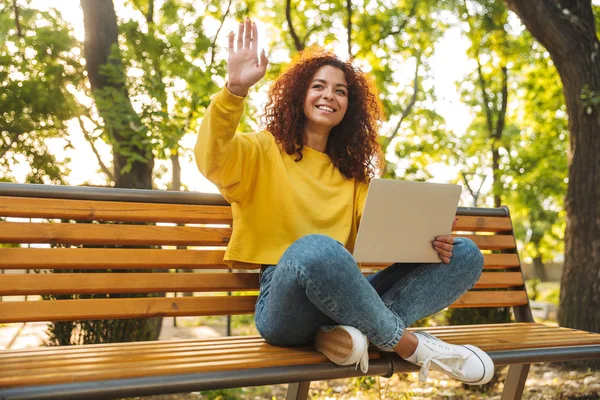 Usmívající se veselou mladou studentkou, která sedí venku v přírodním parku a používá přenosný počítač. — Stock fotografie