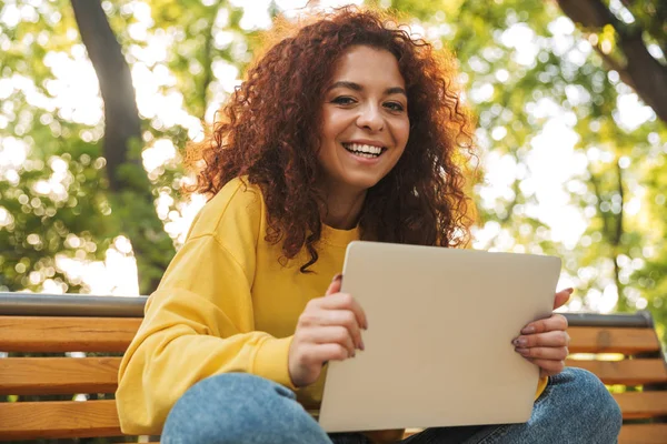 Happy young beautiful curly student girl sitting outdoors in nature park using laptop computer. — Stock Photo, Image