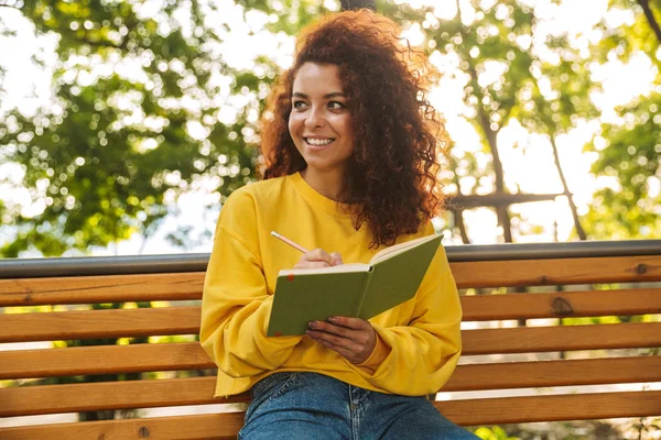 Happy young beautiful curly student girl sitting outdoors in nature park reading book writing notes. — Stock Photo, Image