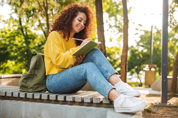 Young beautiful curly student girl sitting outdoors in nature park writing notes in notebook. — Stock Photo, Image