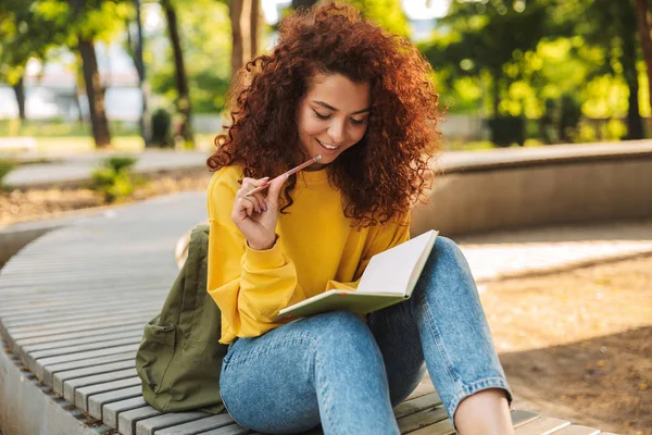 Junge schöne lockige Studentin sitzt draußen im Naturpark und schreibt Notizen in Notizbuch. — Stockfoto