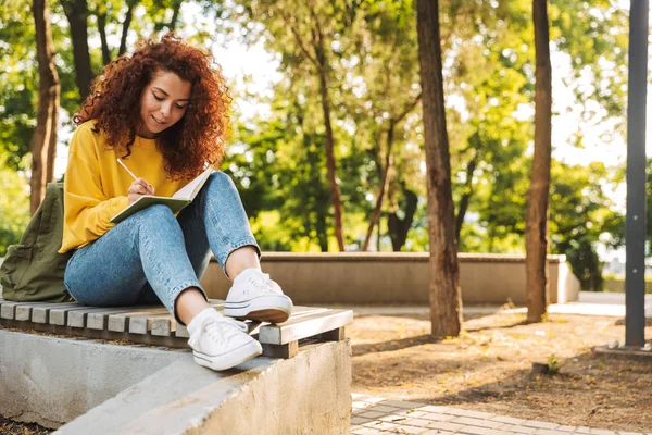 Young beautiful curly student girl sitting outdoors in nature park writing notes in notebook. — Stock Photo, Image