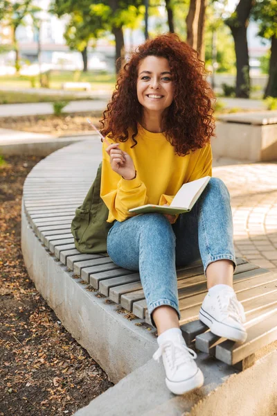 Young beautiful curly student girl sitting outdoors in nature park writing notes in notebook. — Stock Photo, Image