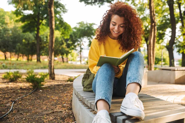 Junge schöne lockige Studentin sitzt draußen im Naturpark und schreibt Notizen in Notizbuch. — Stockfoto