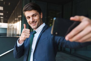 İş merkezinin yakınındaki sokakta açık havada iyimser genç iş adamı cep telefonuyla selfie çekiyor.