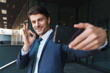 İş merkezinin yakınındaki sokakta açık havada iyimser genç iş adamı cep telefonuyla selfie çekiyor.