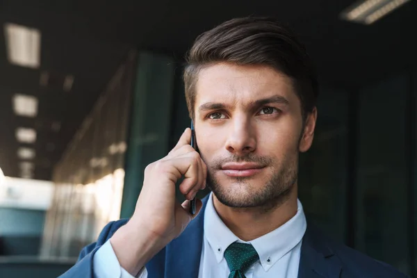 Guapo joven hombre de negocios serio al aire libre en la calle cerca del centro de negocios hablando por teléfono móvil . — Foto de Stock