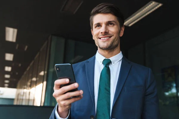 Pleased young business man outdoors at the street near business center using mobile phone. — Stock Photo, Image