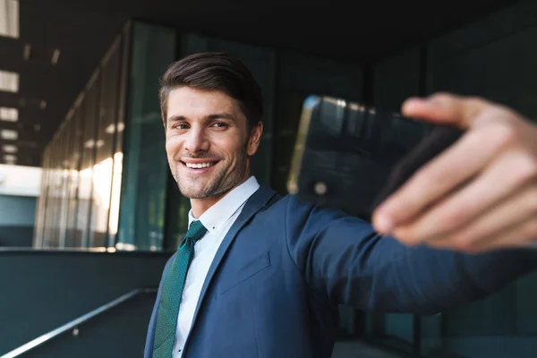 Optimista joven hombre de negocios al aire libre en la calle cerca del centro de negocios tomar selfie por teléfono móvil . — Foto de Stock