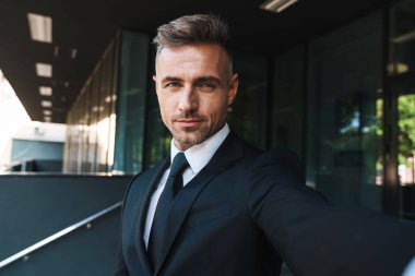 Olgun gri saçlı yakışıklı iş adamı iş merkezi yakınındaki sokakta açık havada kamera ile selfie çekmek.