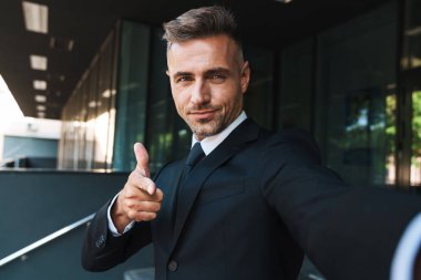 Memnun pozitif olgun gri saçlı yakışıklı iş adamı iş merkezi yakınındaki sokakta açık kamera ile selfie çekmek.