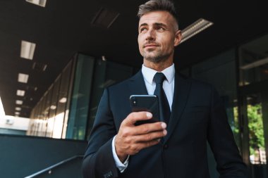 Cep telefonu kullanarak iş merkezi yakınındaki sokakta açık havada gülümseyen olgun gri saçlı yakışıklı iş adamı düşünme.