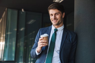 Gülümseyen genç tıraşsız yakışıklı iş adamı açık havada iş merkezi nin yakınındaki sokakta kahve içiyor