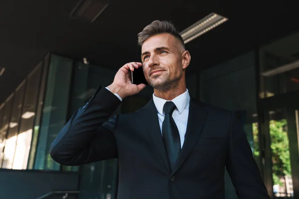 Оптимістичний зрілий сіроокий красивий бізнесмен на вулиці біля бізнес-центру розмовляє по мобільному телефону . — стокове фото