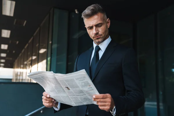 Hombre de negocios maduro guapo de pelo gris concentrado al aire libre en la calle cerca del centro de negocios leyendo el periódico . — Foto de Stock