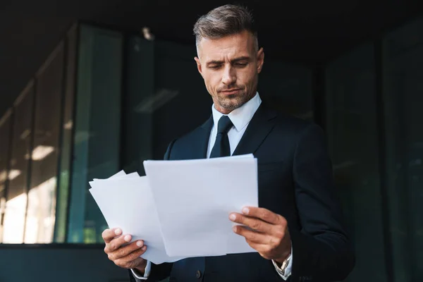 Sérieux mature beau aux cheveux gris homme d'affaires à l'extérieur dans la rue près du centre d'affaires détenant des documents . — Photo