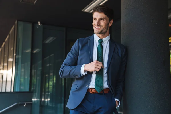 Cheerful happy young unshaved handsome business man outdoors at the street near business center posing touching tie. — Stock Photo, Image