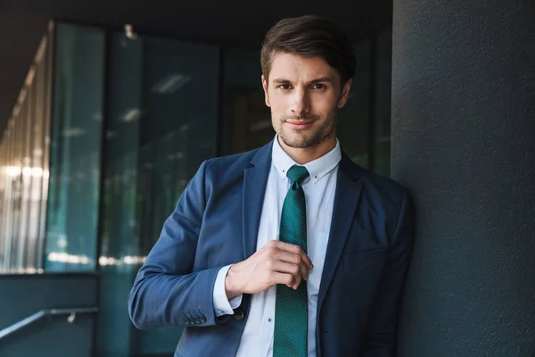 Pleased young unshaved handsome business man outdoors at the street near business center posing touching tie. — Stock Photo, Image