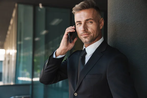 Sérieux mature beau aux cheveux gris homme d'affaires à l'extérieur dans la rue près du centre d'affaires parler par téléphone mobile . — Photo