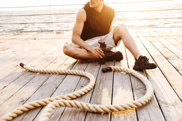 Foto di uomo non rasato in tuta da ginnastica che riposa e beve acqua mentre si allena con corda fitness sul molo di legno al mare — Foto Stock