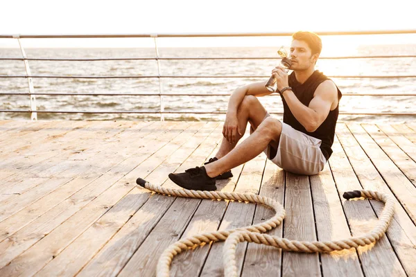 Foto del giovane in tuta da ginnastica che riposa e beve acqua mentre si allena con la corda da fitness sul molo di legno al mare — Foto Stock