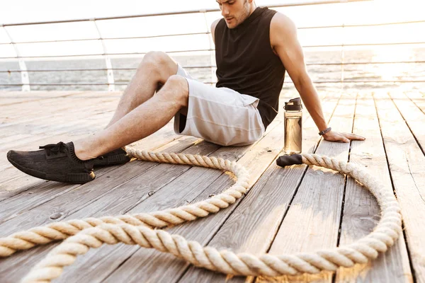 Foto de un hombre fuerte de 20 años en chándal descansando mientras hace ejercicio con una cuerda de fitness en un muelle de madera en la playa — Foto de Stock